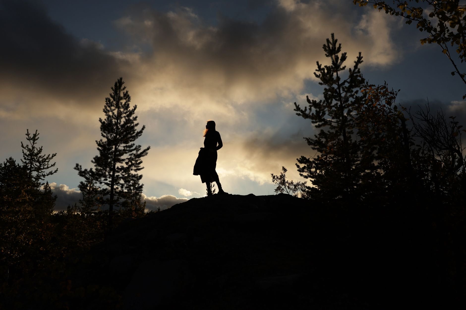 A woman stands silhouetted against a dramatic sunset sky surrounded by trees.